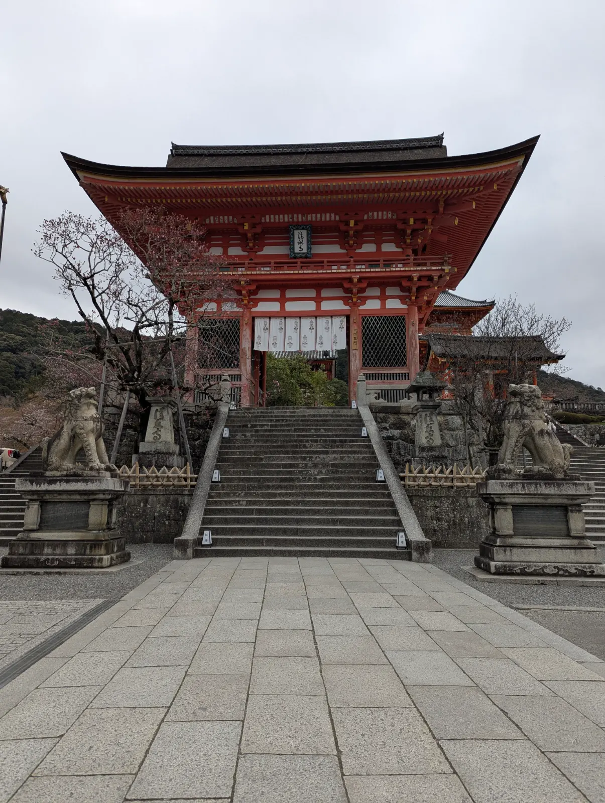 The iconic vibrant orange-red gate of Kiyomizu-dera Temple in Kyoto, Japan, with stone lion statues guarding the grand staircase leading up to the temple.