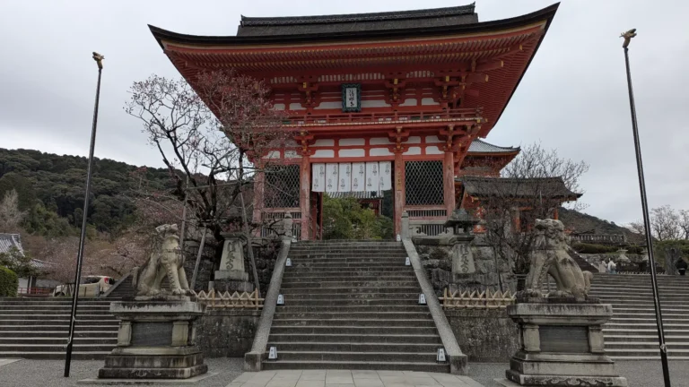 The grand vermilion gate of Kiyomizu-dera stands at the top of the stone steps, flanked by guardian lion-dogs, beneath a grey Kyoto sky, a sacred threshold between city and stillness.