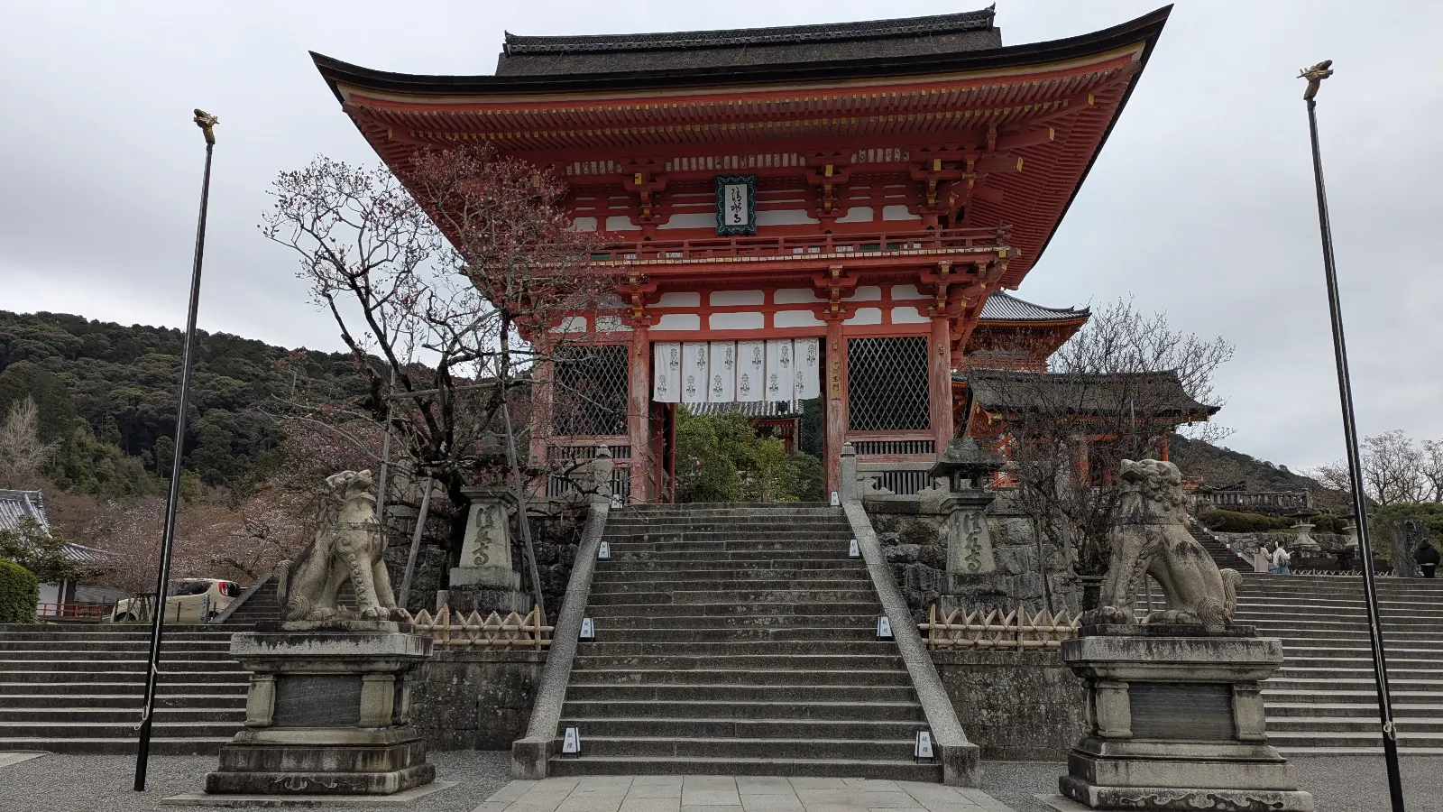 The grand vermilion gate of Kiyomizu-dera stands at the top of the stone steps, flanked by guardian lion-dogs, beneath a grey Kyoto sky, a sacred threshold between city and stillness.