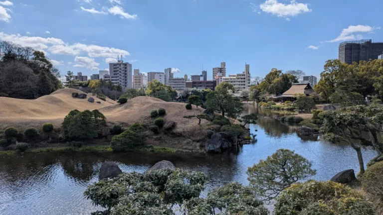 Off the beaten path Kumamoto: Scenic view of Suizenji Jojuen Garden with a backdrop of the city skyline, blending nature’s tranquility with the urban setting.