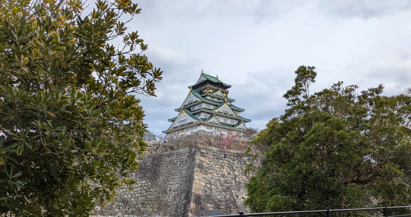 Osaka Castle rising above stone walls and green trees, under a soft spring sky.